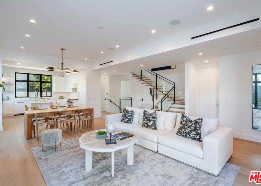 Modern open-concept living room and kitchen with white furnishings, wood accents, a dining table, and a staircase leading upstairs.