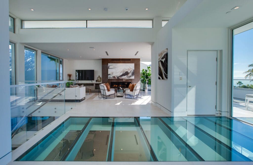 Modern living room with glass floor panels, white walls, large windows, a fireplace, and contemporary furniture. Ocean view visible through the windows.