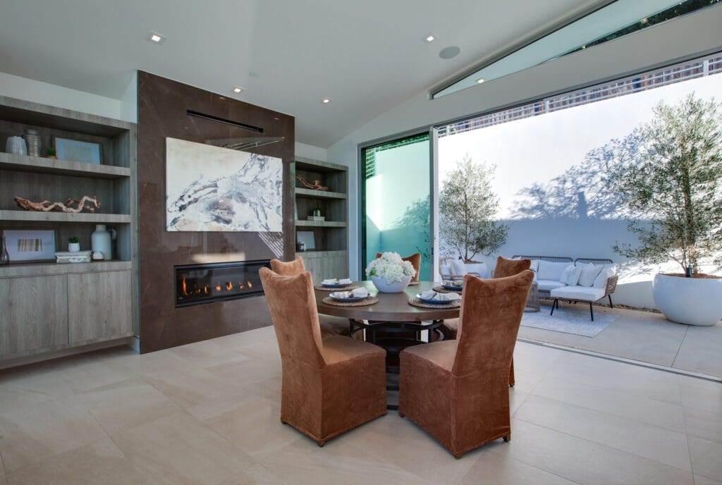 Modern indoor dining area with a round table and four brown chairs near a fireplace, opening to an outdoor patio with seating and potted trees.