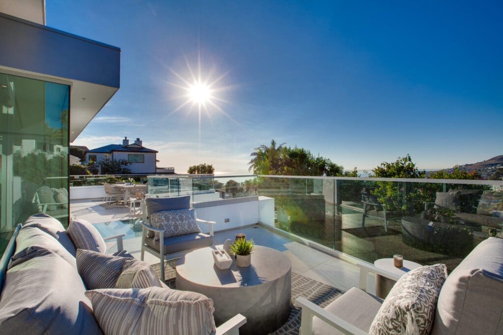 Modern outdoor patio with cushioned seating, a round coffee table, and glass railing overlooks a sunny landscape with clear blue sky and distant ocean view.