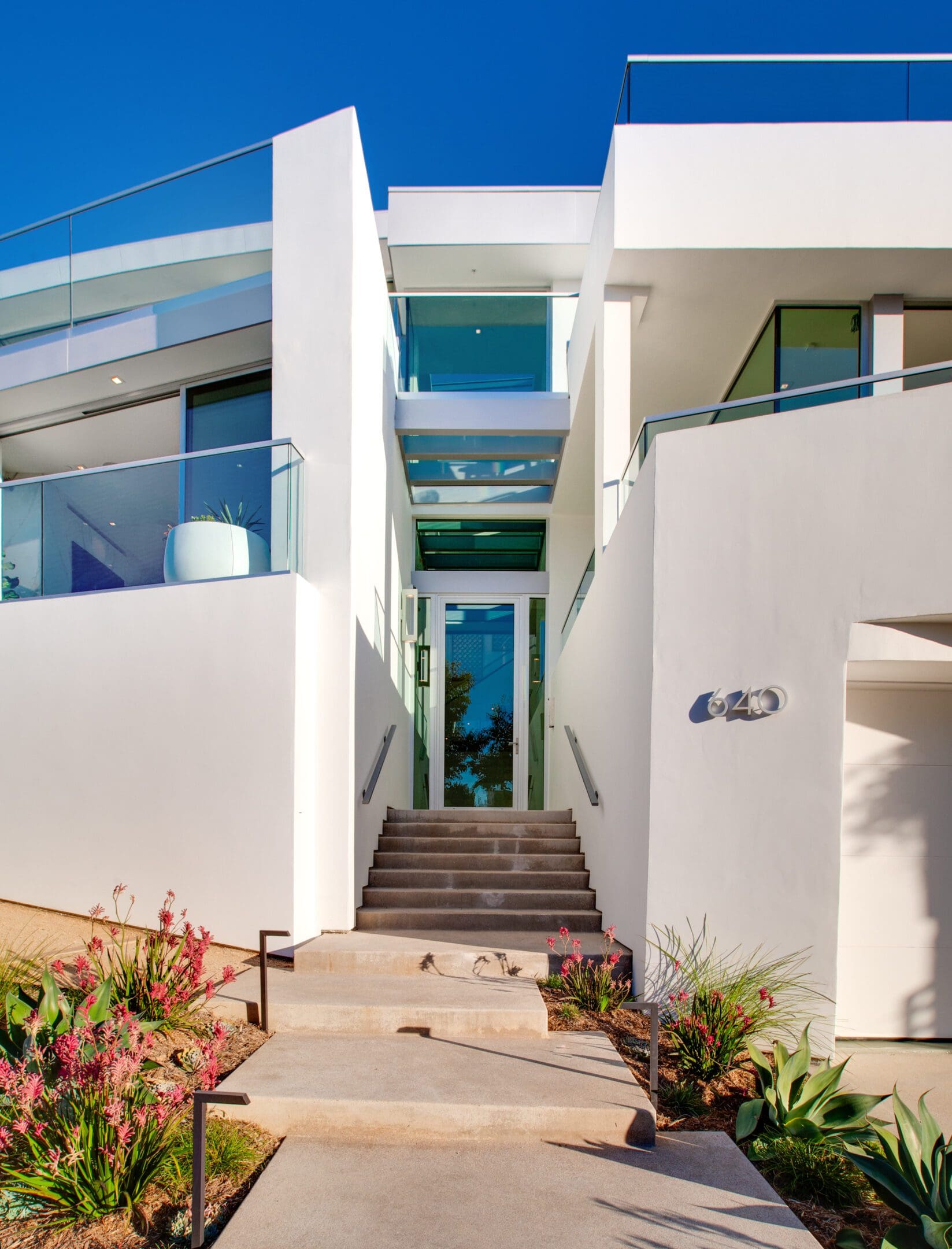 Modern white two-story house with glass railings, large windows, and central entrance, surrounded by manicured landscaping under a clear blue sky.