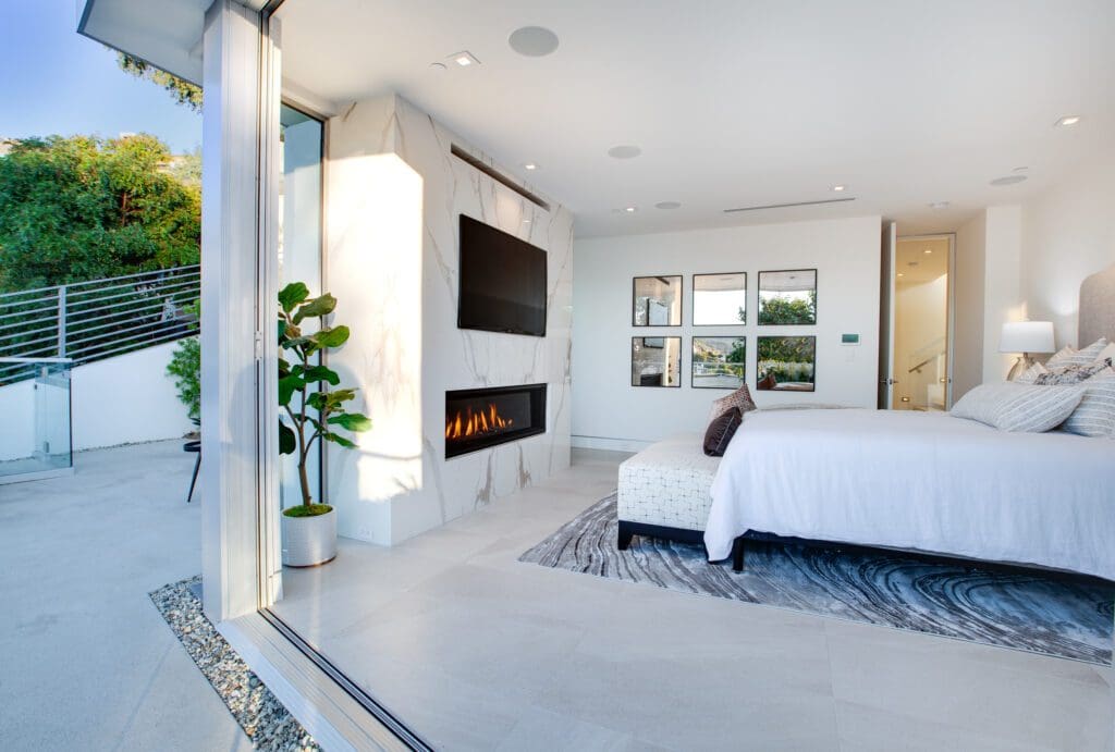 Modern bedroom with open glass doors to a patio, white walls, a bed with white bedding, a fireplace under a wall-mounted TV, and a tall plant in the corner.
