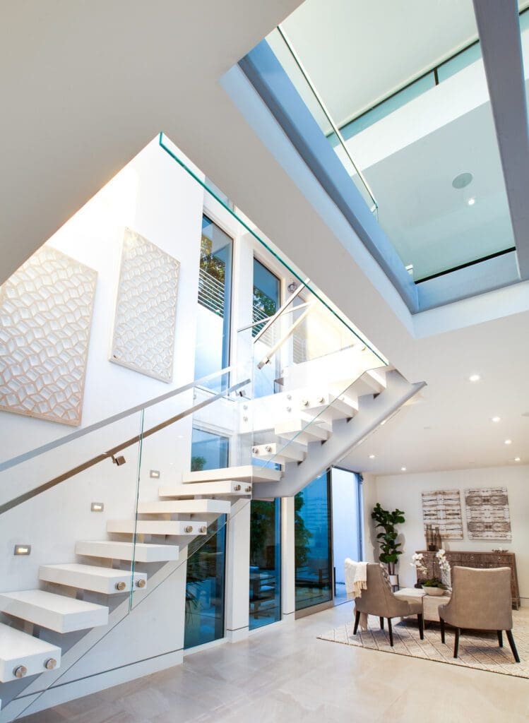 Modern interior with floating staircase, glass railing, tall windows, and skylight. Dining area with two chairs and table is visible below. Neutral color palette throughout.