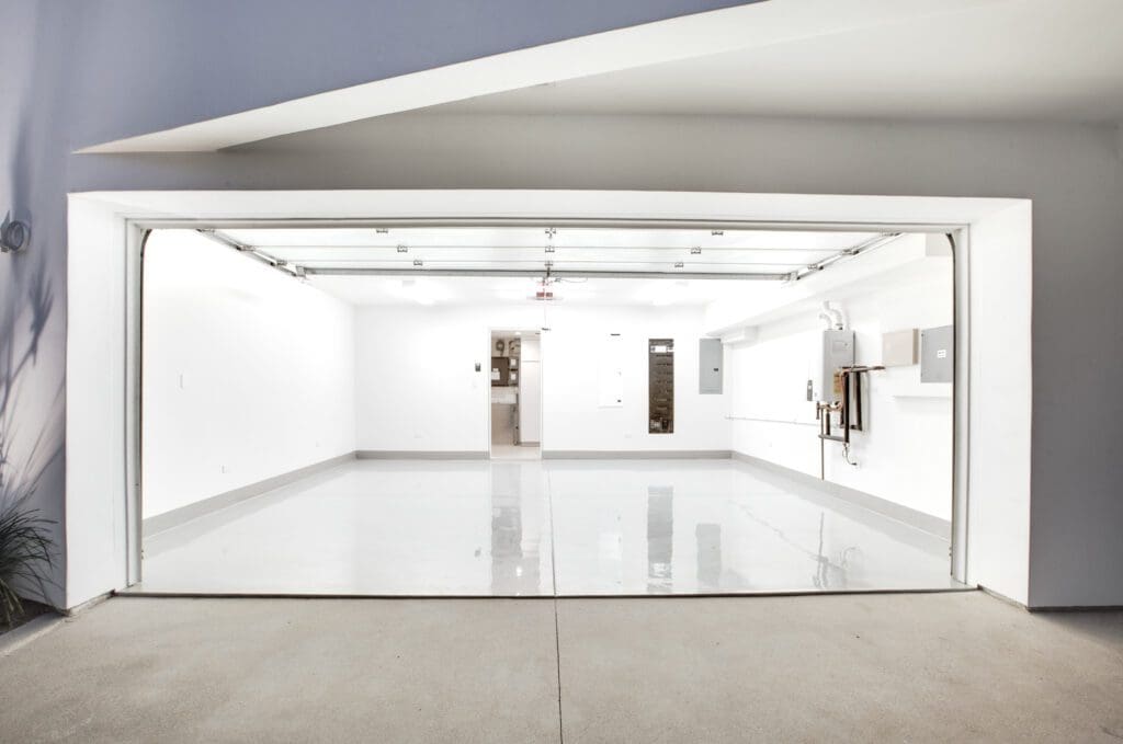 A clean, empty, modern garage with white walls, a glossy floor, and an open garage door facing outward. Electrical panels and pipes are mounted on the right wall.