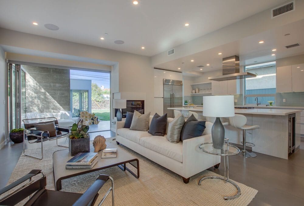 Modern open-concept living room and kitchen with neutral tones, a white sofa, bar stools, and large sliding glass doors opening to an outdoor patio.
