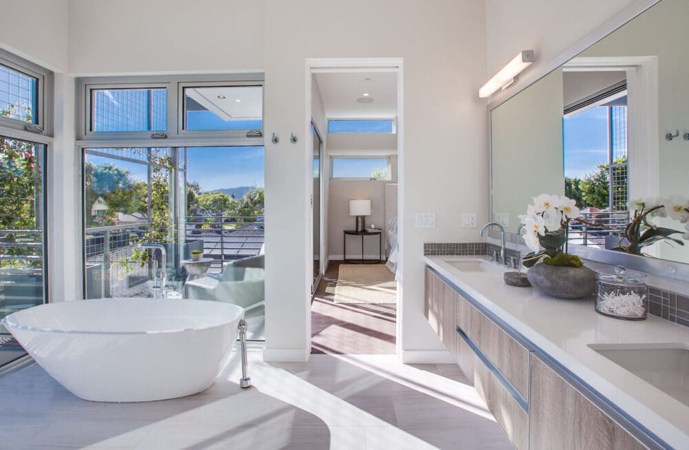Modern bathroom with a freestanding bathtub, double vanity, large mirror, and floor-to-ceiling windows overlooking a balcony; bedroom visible through open doorway.