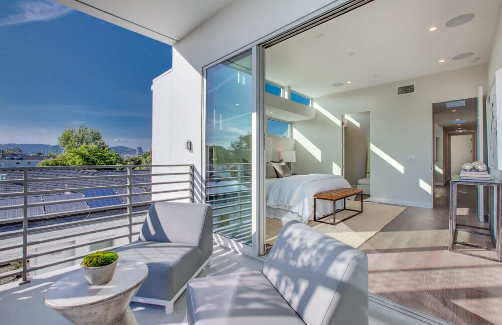 Modern bedroom with open sliding glass doors leading to a balcony with two chairs and a small table, overlooking rooftops with solar panels and greenery under a clear sky.