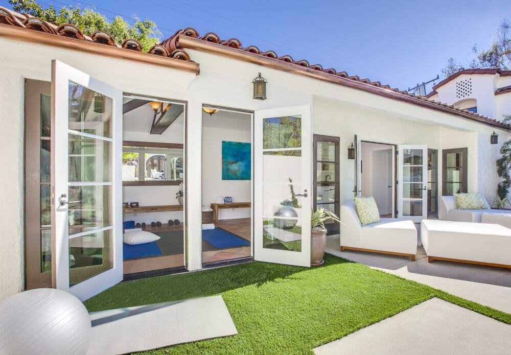 Open glass doors lead from a bright yoga studio to a patio with artificial grass, white outdoor furniture, and potted plants in a sunny backyard.