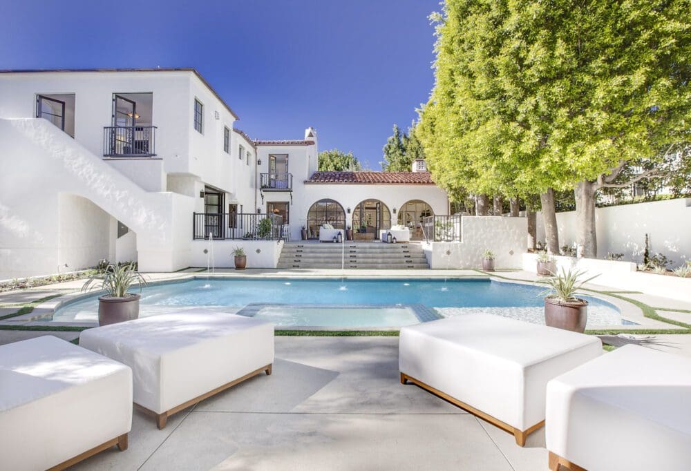Luxurious white villa with arched patio, outdoor pool, sun loungers, potted plants, and manicured trees under a clear blue sky.