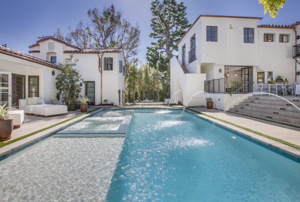 Modern backyard with large swimming pool, sun loungers, white two-story house, water fountains, and landscaped greenery under a clear blue sky.