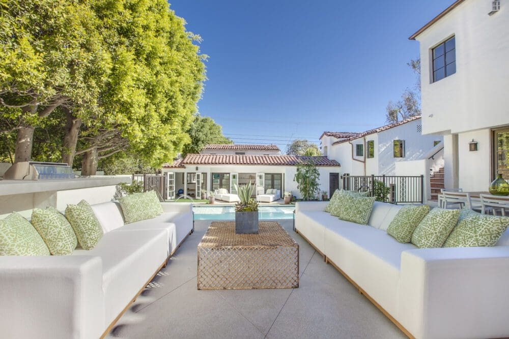 Outdoor patio seating area with white sofas and green pillows, a central coffee table, and a view of a pool and white house with red tile roof in the background on a sunny day.