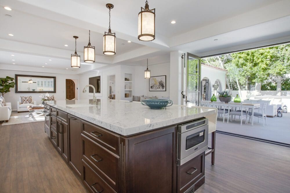 Modern kitchen with a large island, marble countertop, built-in appliances, pendant lights, and open sliding doors leading to an outdoor dining area.