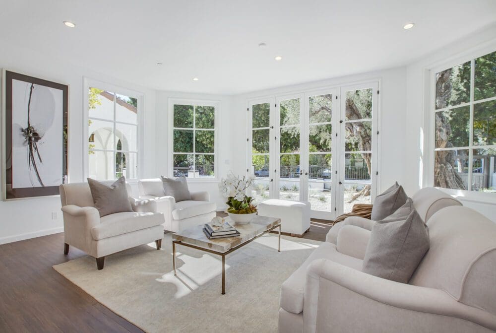 A bright living room with large windows, white armchairs, a beige sofa, glass coffee table, and neutral decor on a light rug over hardwood floors.