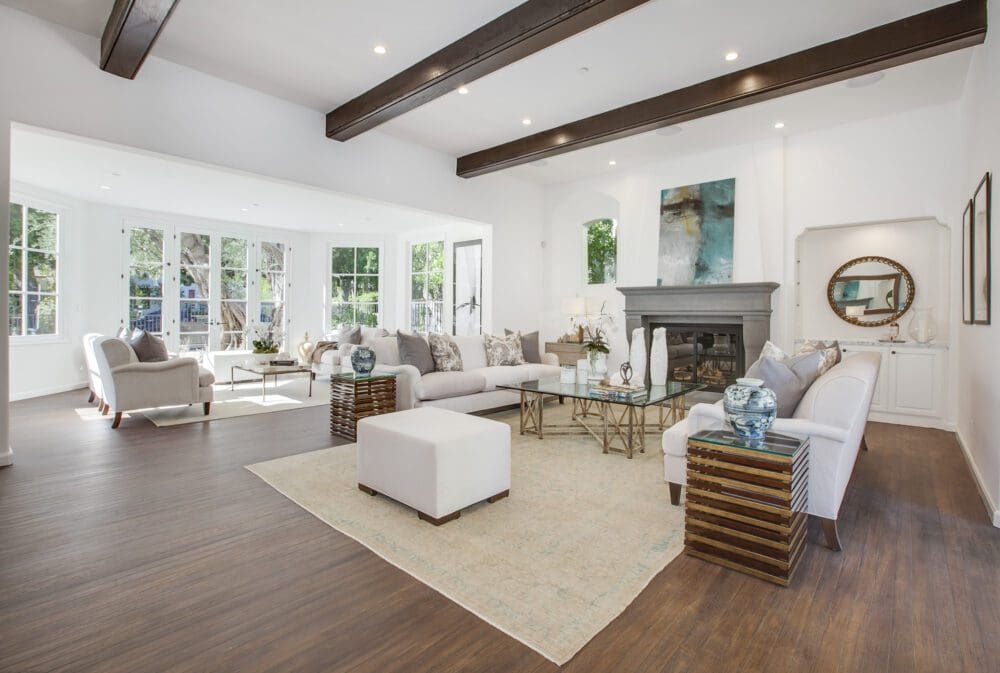 Spacious, modern living room with wood floors, exposed ceiling beams, large windows, neutral furniture, a glass coffee table, fireplace, and contemporary decor.