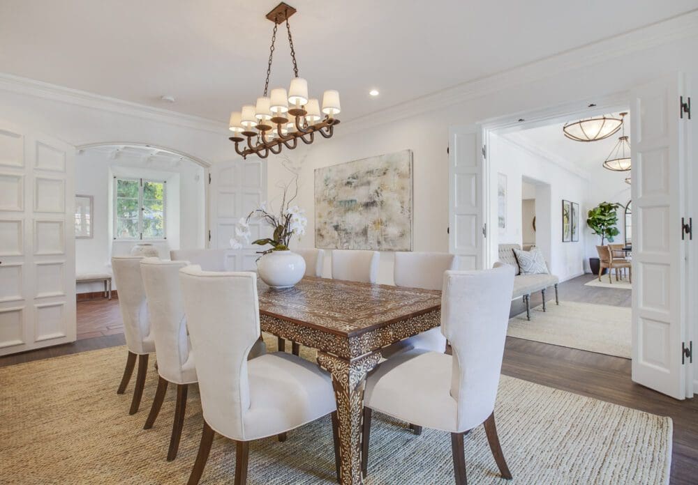A dining room with a carved wooden table, eight white upholstered chairs, a chandelier, beige rugs, and abstract wall art. French doors lead to a bright adjoining room.