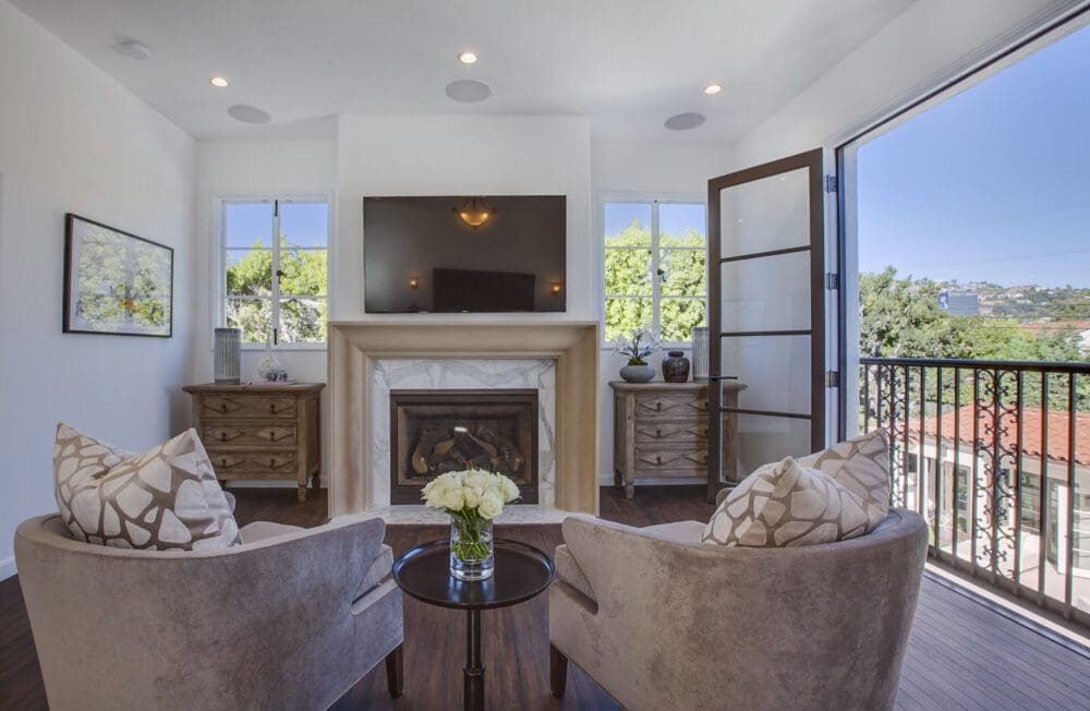A modern living room with two armchairs, a round table with flowers, a fireplace, a wall-mounted TV, and double doors opening to a balcony with a view of trees and rooftops.
