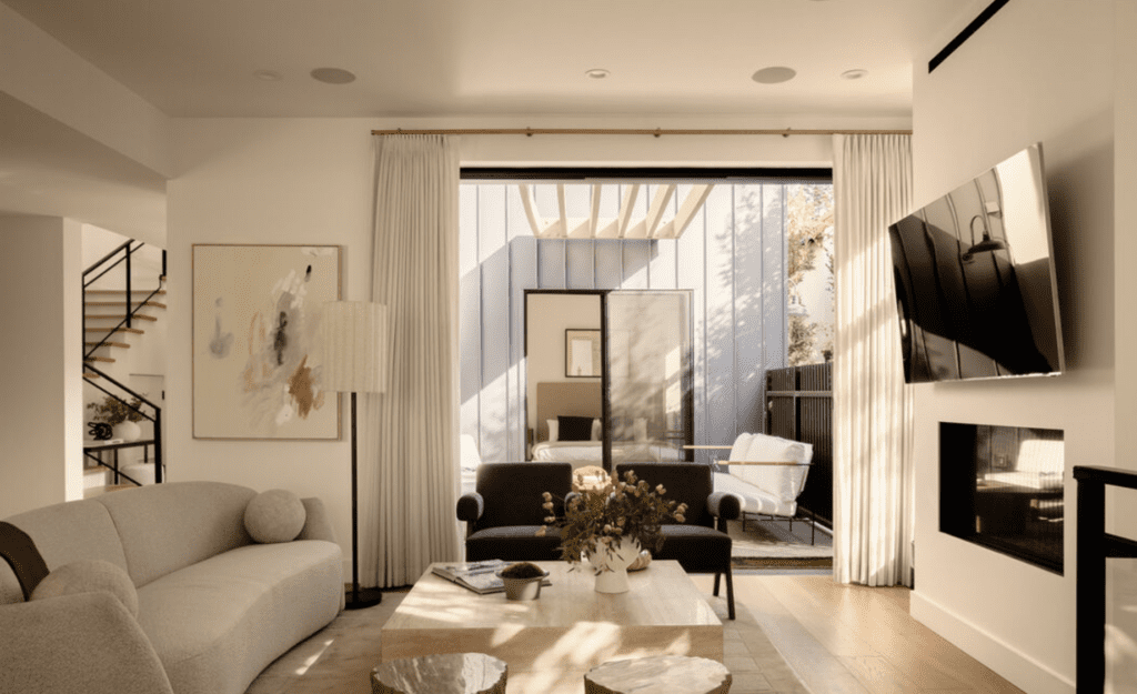 Bright modern living room with neutral furniture, a wall-mounted TV, abstract art, and large glass doors opening to a patio with outdoor seating.