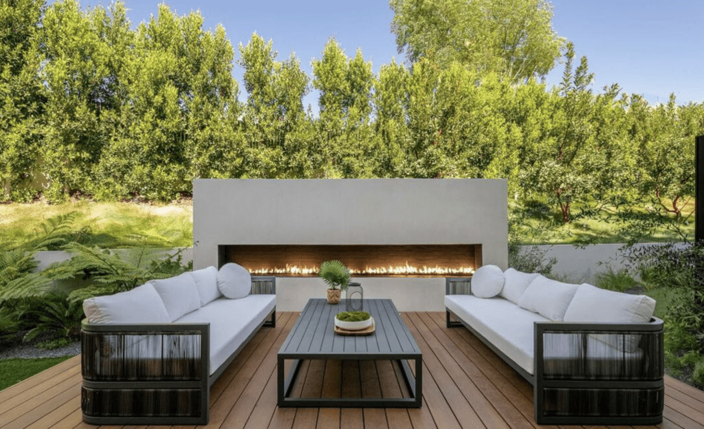 Two modern outdoor sofas with white cushions face each other across a black coffee table on a wooden deck, with a built-in fireplace and green hedges in the background.