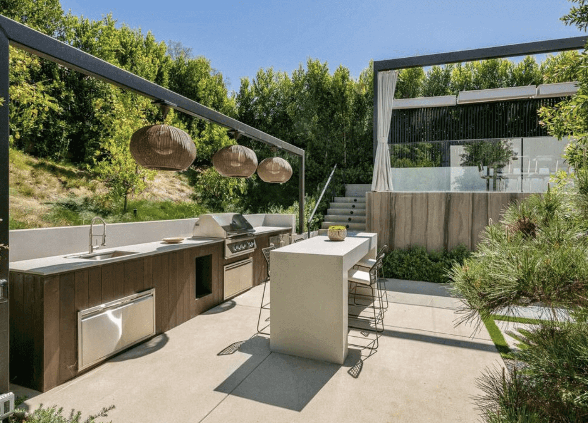 Modern outdoor kitchen with a grill, sink, island seating, and pendant lights, surrounded by greenery and a raised deck with glass railing in the background.