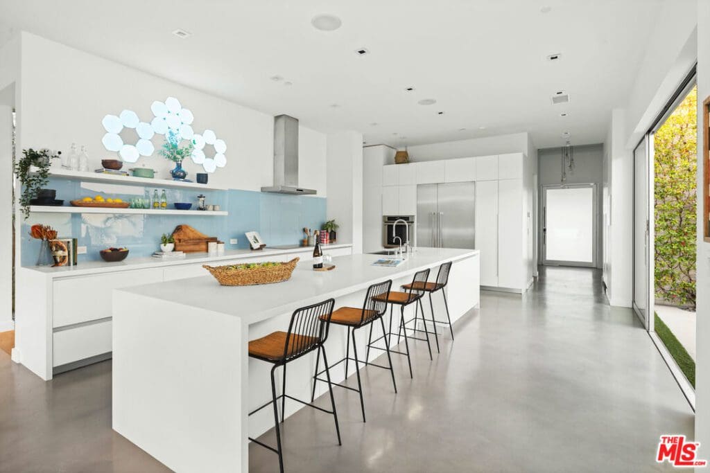 Modern white kitchen with a large island, four bar stools, open shelving, stainless steel appliances, and glass door leading outside.