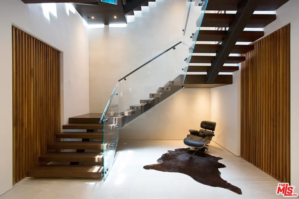 A modern interior features a floating wooden staircase with glass railings, wooden slat walls, and a lounge chair with an ottoman on a dark cowhide rug.