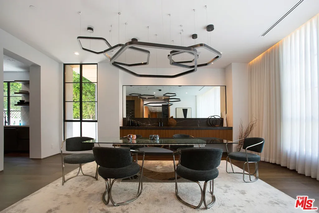 Modern dining room with a glass table, six dark chairs, geometric chandelier, large mirror, and floor-to-ceiling curtains.