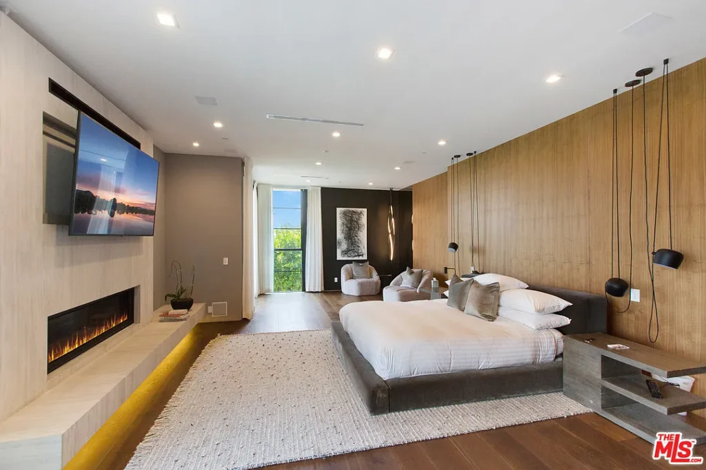 Modern bedroom with a large bed, wall-mounted TV, linear fireplace, seating area, and floor-to-ceiling windows letting in natural light.