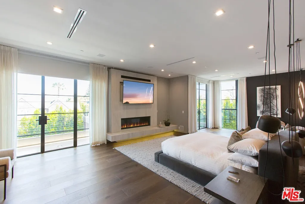 Modern bedroom with a large bed, wall-mounted TV above a fireplace, floor-to-ceiling windows, dark wood floors, and neutral decor.