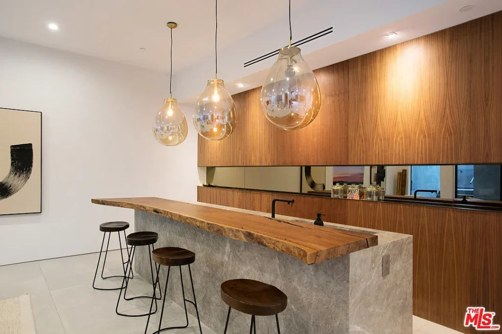 Modern kitchen with a live-edge wooden island, four metal stools, pendant lights, and wood-paneled cabinetry with a mirrored backsplash.