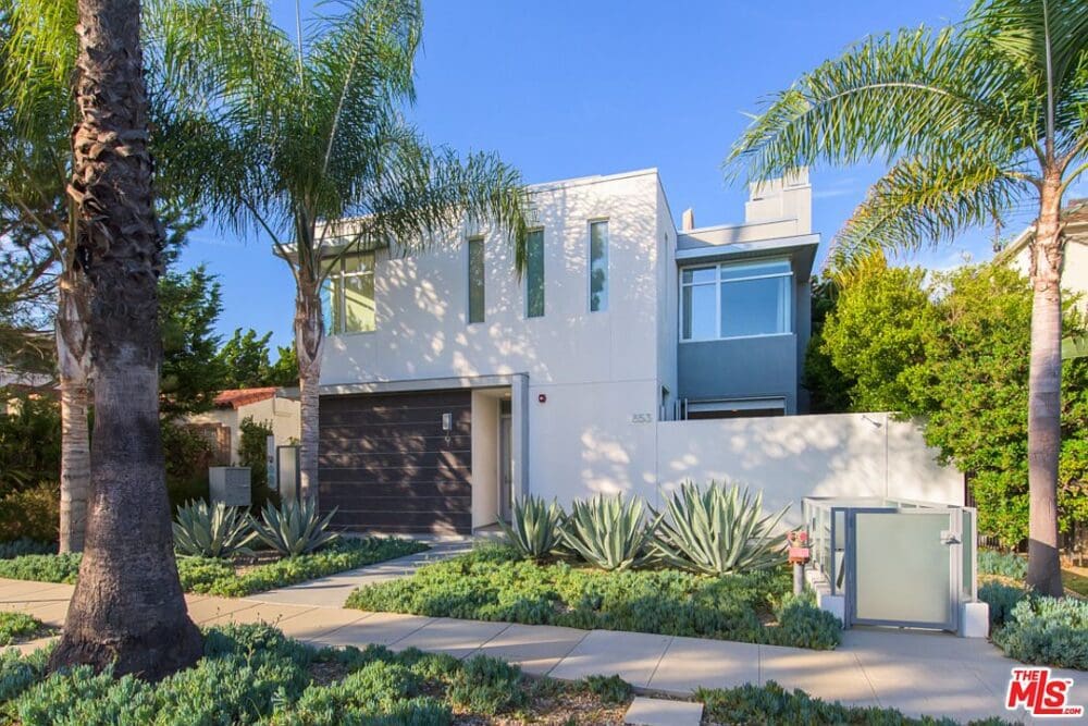 Modern two-story house with a flat roof, large windows, and a mix of white and blue-gray exterior, surrounded by palm trees and desert landscaping on a sunny day.