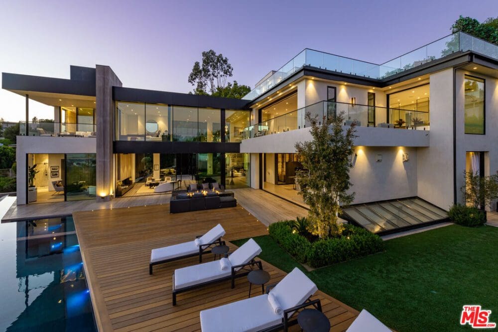 Modern luxury home with large glass windows, spacious wooden deck, pool, outdoor lounge, and several lounge chairs at dusk.