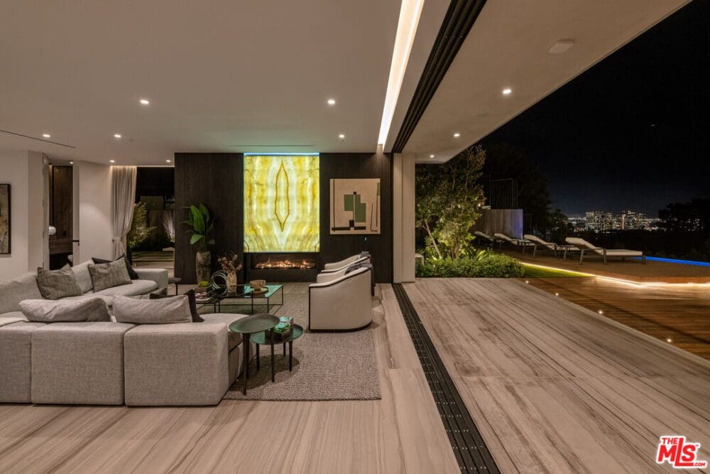 Modern open-plan living room with neutral furnishings, illuminated fireplace, large sliding doors, and city skyline view at night. Outdoor seating and pool area are visible.