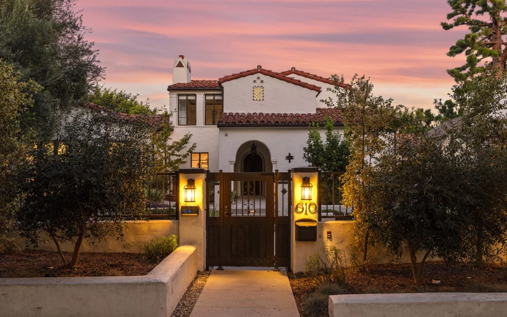 A two-story Spanish-style house with a red tile roof, arched doorway, and illuminated exterior lights, viewed at sunset behind a wooden gate marked “610.”.