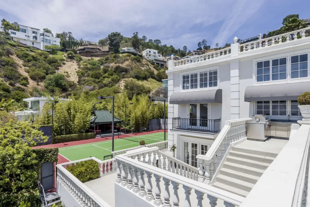 A large white house with balconies overlooks a fenced tennis court, surrounded by hills and greenery under a partly cloudy sky.