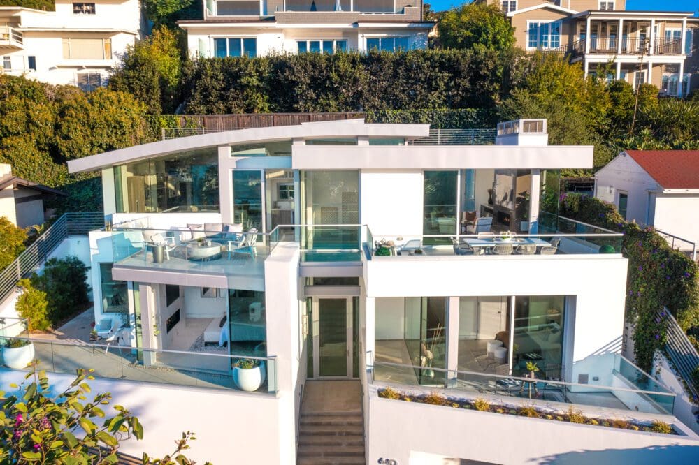 Modern white multi-level house with large glass windows, multiple balconies, and outdoor seating areas, surrounded by greenery and neighboring homes.