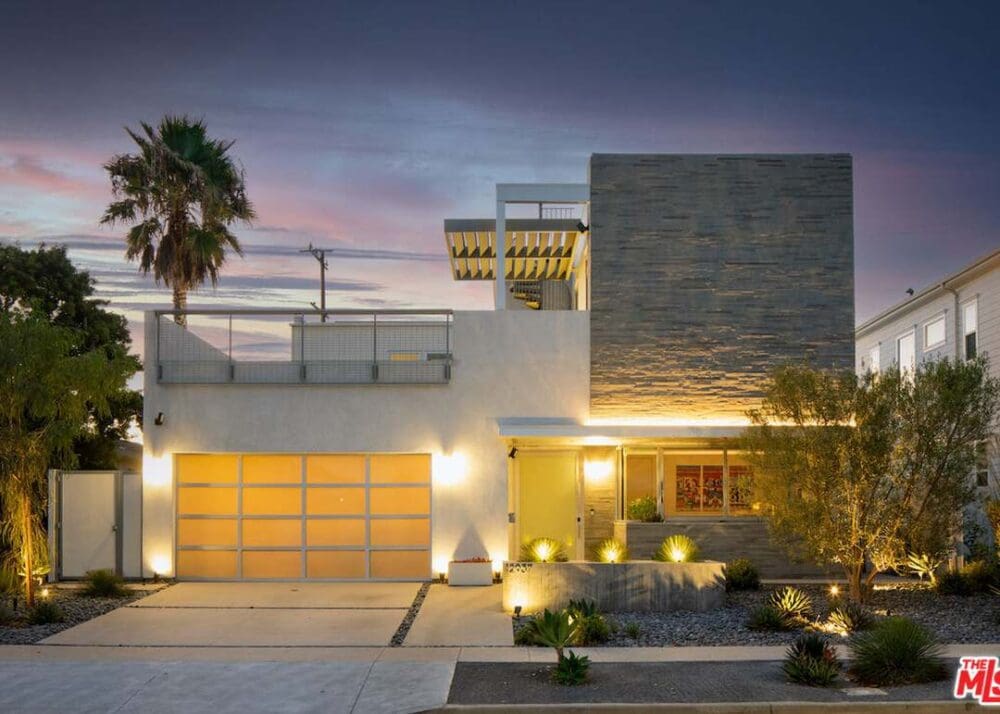 Modern two-story house at dusk with flat roof, stone facade, large glass garage door, rooftop terrace, and landscaped front yard with accent lighting.