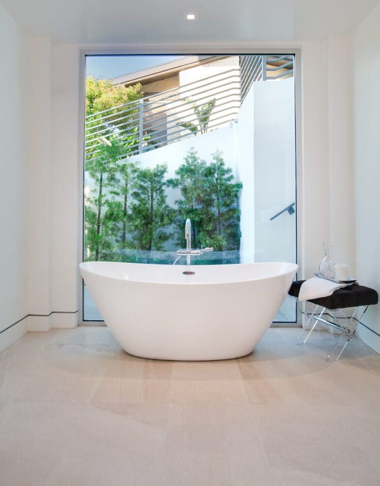 A modern white bathtub is centered in front of a large window with a view of greenery and a building exterior; a small stool with towels sits to the side.