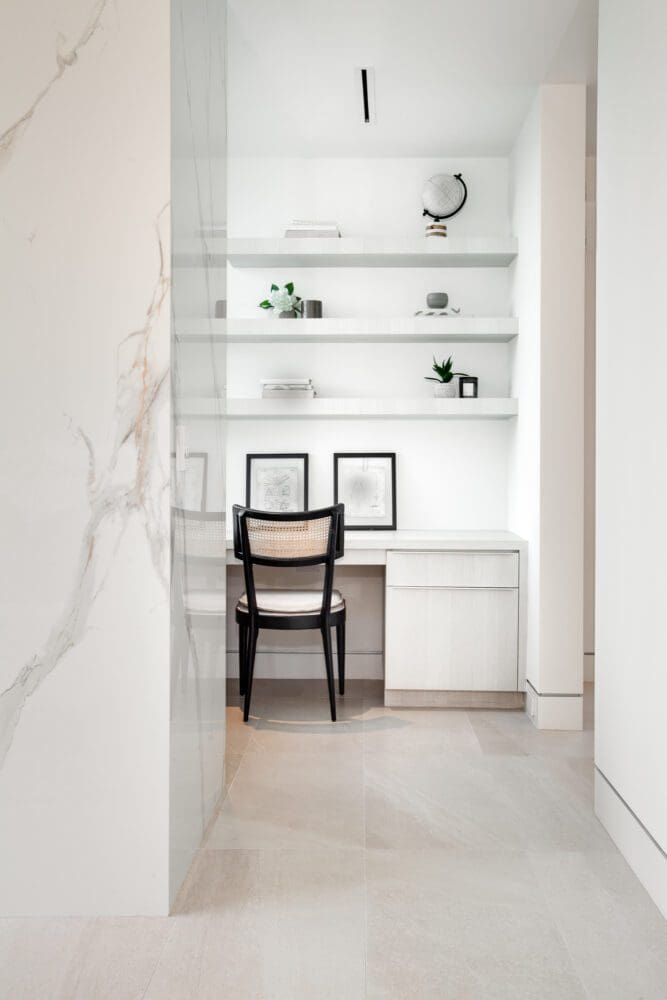 A minimalist home office with white shelves, a small desk, two framed artworks, a chair, and decorative items against a white wall.