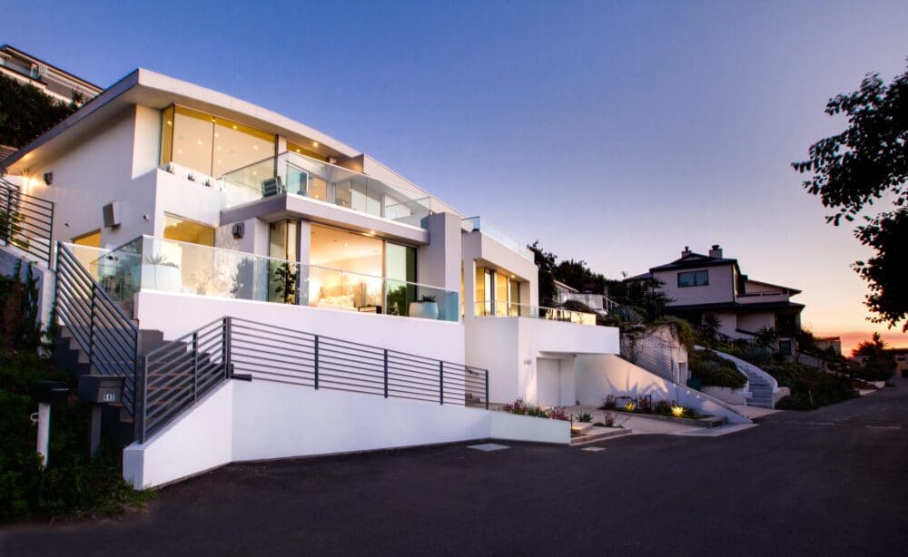 Modern two-story house with large glass windows, balconies, and white exterior, located on a sloped street at sunset.