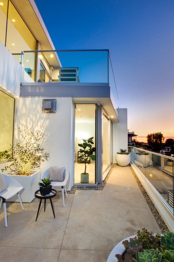 Modern apartment balcony at sunset with glass railings, potted plants, and outdoor seating, adjacent to large windows and an open sliding glass door.