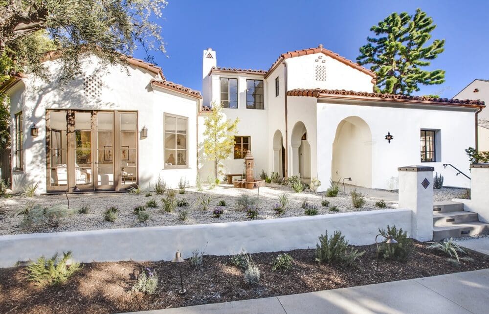 White stucco house with red tile roof, arched entryway, and a minimalist garden in front, set against a clear blue sky.