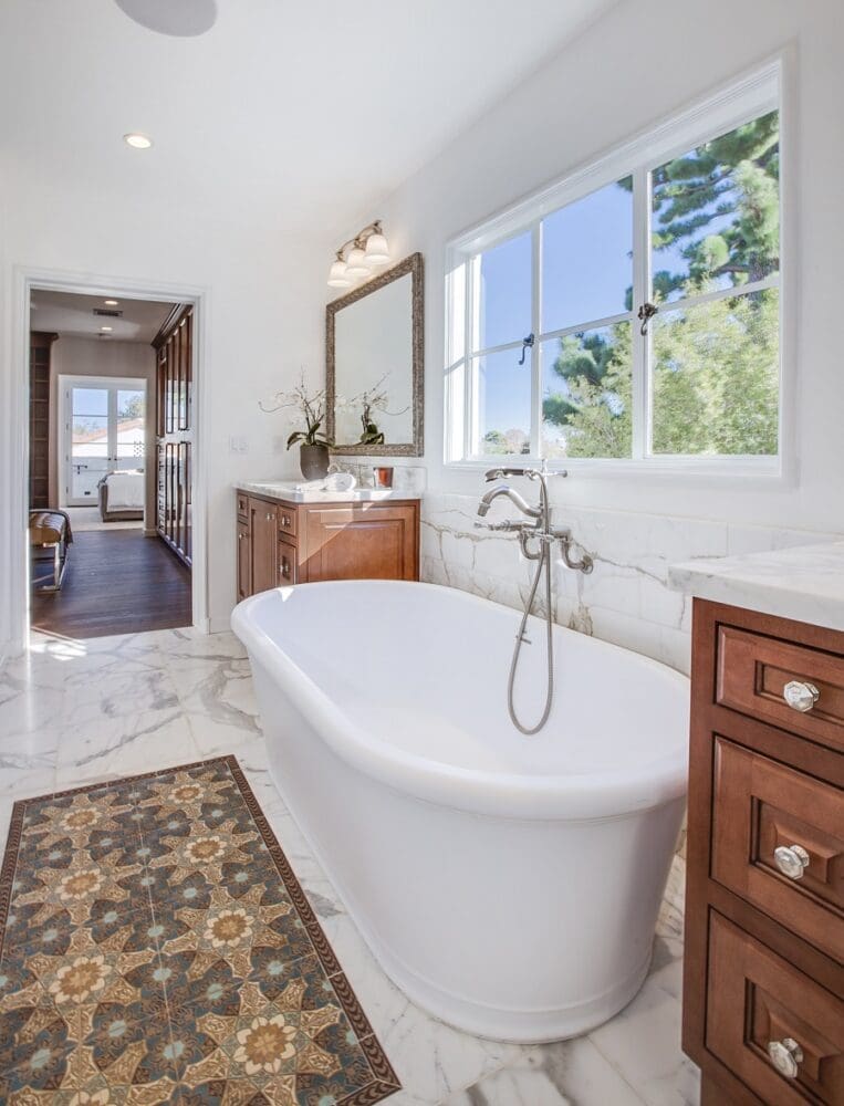 Bright bathroom with a white freestanding tub, marble floor, wooden cabinets, large window, wall mirror, and a patterned rug.