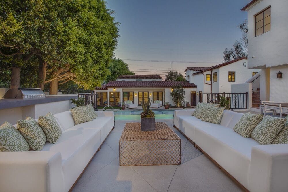 Outdoor patio with white cushioned sofas and patterned pillows, centered table, and view of a swimming pool and Spanish-style house in the background at dusk.