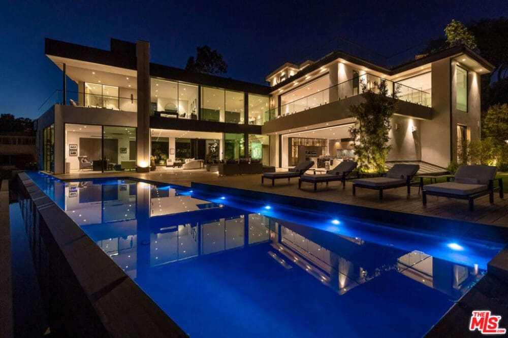 Modern multi-story house at night with large glass windows, outdoor lounge chairs, illuminated pool, and landscaped surroundings.