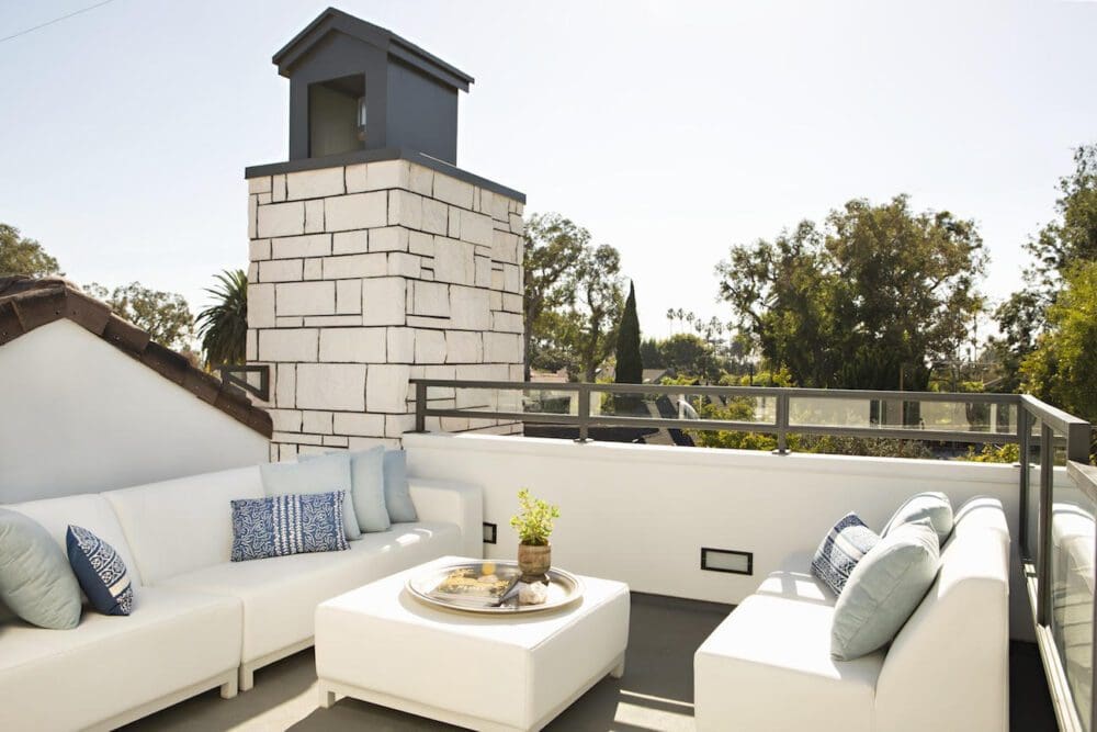 Modern rooftop patio with white sectional sofas, a square coffee table with refreshments, and a stone chimney; trees and houses visible in the background.