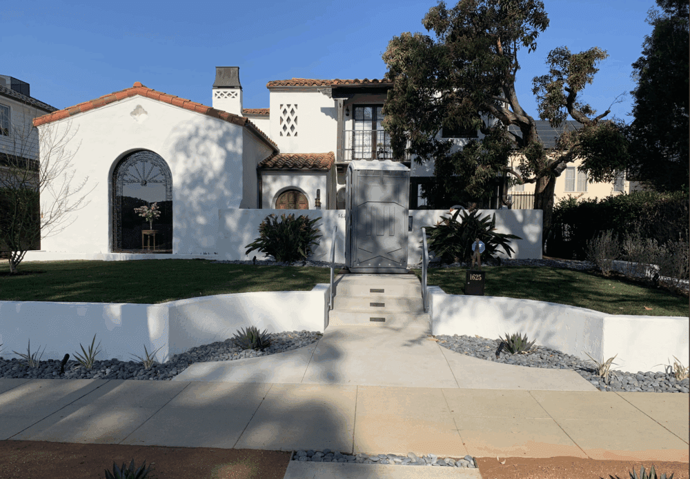 Two-story white stucco house with red tile roof, arched windows, and a landscaped front yard with a short stairway leading to the front door.