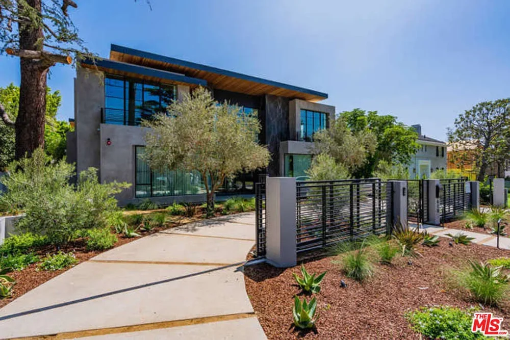 Modern two-story house with large windows, surrounded by landscaped garden and trees, with a metal gate and concrete walkway leading to the entrance.