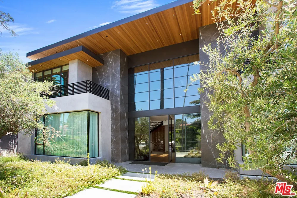 Modern two-story house with large glass windows, dark exterior accents, wood-paneled roof overhang, and landscaped front garden under a sunny blue sky.