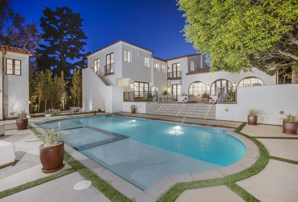 A modern two-story white house with large windows, outdoor stairs, and a swimming pool with spa in a landscaped backyard at dusk.